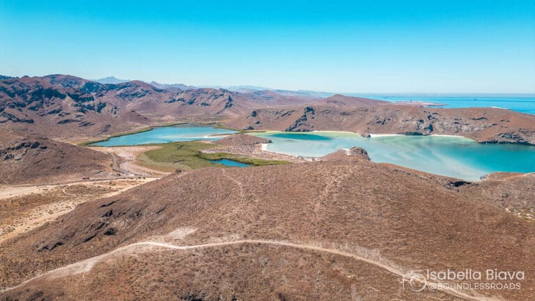 Aerial view of a serene turquoise bay surrounded by arid desert hills and rocky mountains, under a clear blue sky.