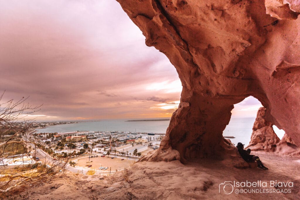A person sits in a rocky cave overlooking a coastal cityscape and marina under a dramatic, colorful sky at sunset.