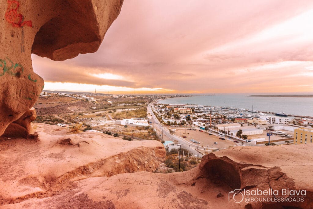 A coastal town view from a rocky hill, overlooking a marina and buildings under a dramatic sunset sky.