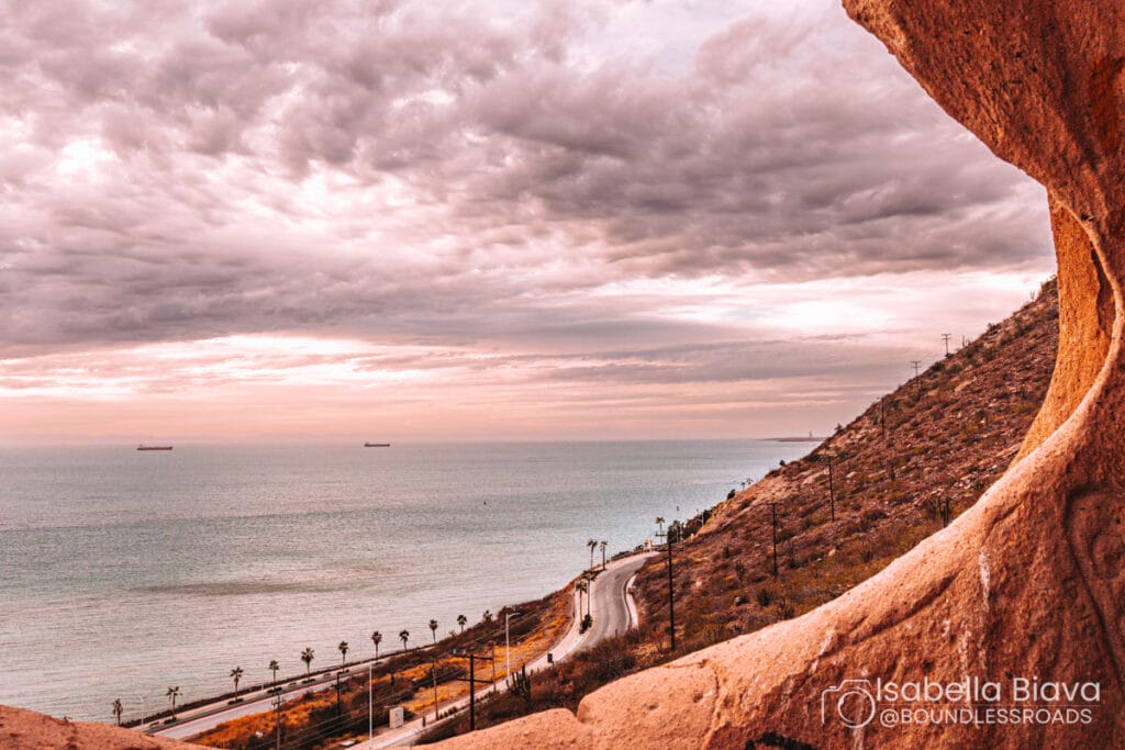 Curved coastline with ocean view, road curves alongside rocky hills, palm trees scattered. Cloudy sky with two cargo ships in the distance.