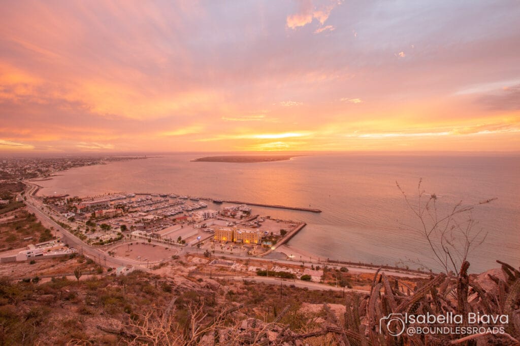 Sunset view over La Paz Bay, Mexico, with a vibrant sky, marina, and distant cityscape, surrounded by the calm sea and rugged coastline.