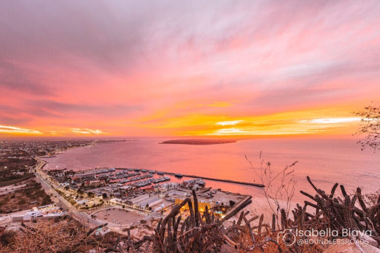 Stunning sunset view over La Paz Bay, Mexico, with vibrant pink and orange skies, marina, and cityscape framed by foreground cacti.
