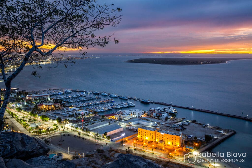 A scenic bay view at sunset, featuring a marina with numerous boats, illuminated buildings, and vibrant city lights under a colorful sky.