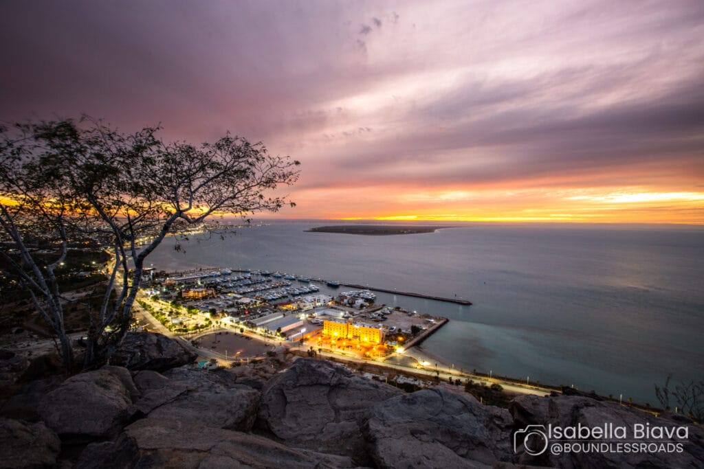 A vibrant sunset over Marina La Paz, Mexico, with illuminated streets and distant islands, viewed from a rocky hillside with silhouetted trees.